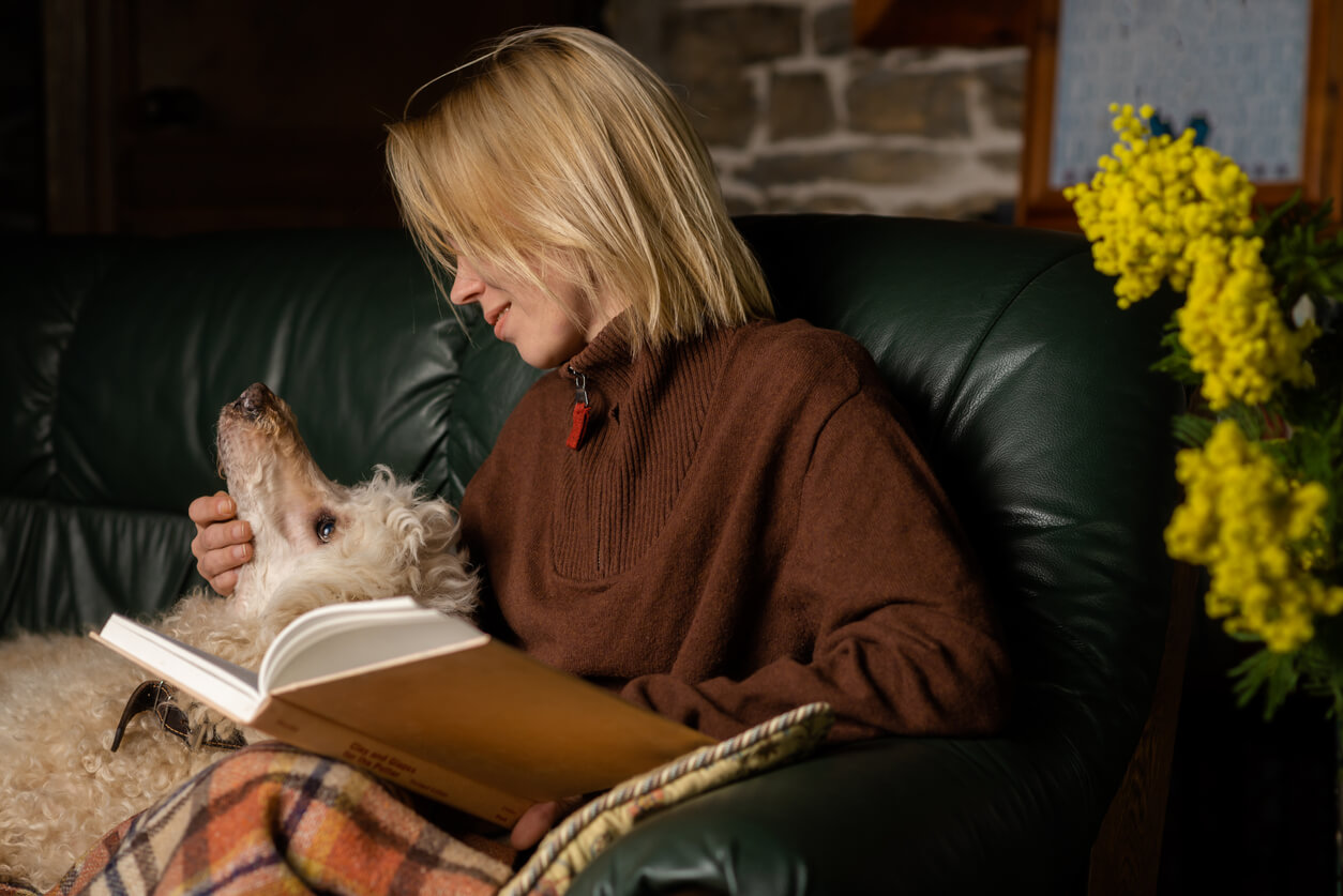 Mujer acaricia a su perro, mientras lee un libro en el sofá. 
